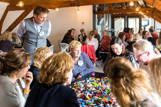 Deelnemers verspreid over meerdere tafels tijdens een evenement met LEGO® bouwstenen met Marcel de Jong die toekijkt hoe ze bouwen